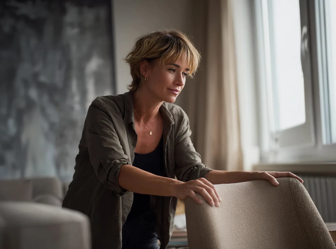 A woman pausing thoughtfully beside an upholstered armchair in a softly lit living room